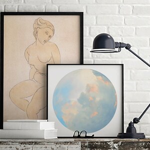 May include: Two framed prints on a white brick wall. The print on the left is a line drawing of a nude woman. The print on the right is a painting of a blue and pink cloudy sky.