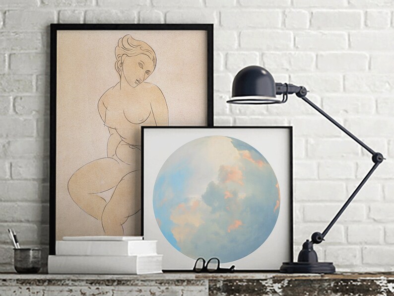 May include: Two framed prints on a white brick wall. The print on the left is a line drawing of a nude woman. The print on the right is a painting of a blue and pink cloudy sky.