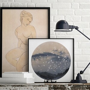 May include: Two framed prints on a white brick wall. The print on the left is a line drawing of a nude woman. The print on the right is a circular painting of a sky with white birds flying over a dark blue cloud.