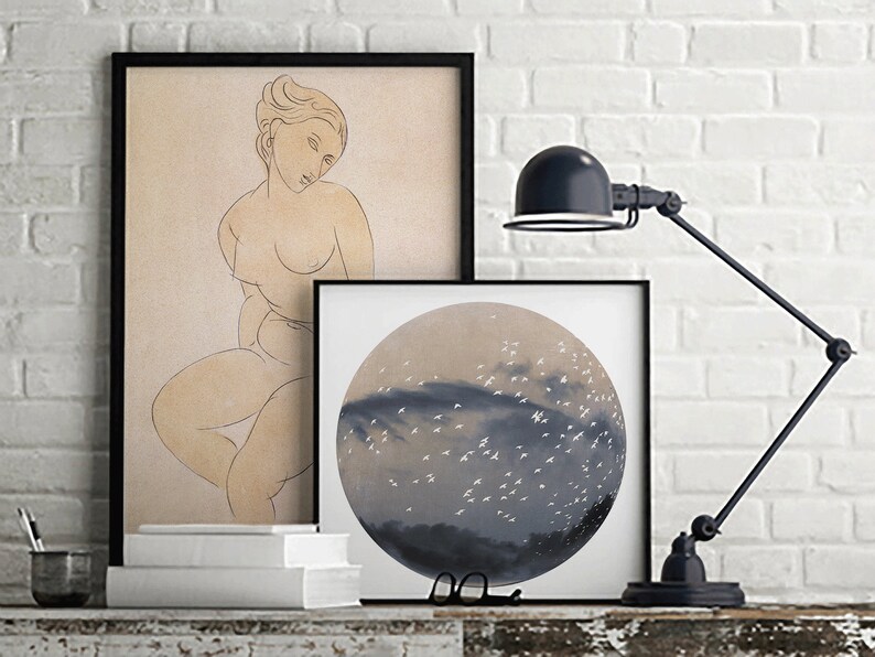 May include: Two framed prints on a white brick wall. The print on the left is a line drawing of a nude woman. The print on the right is a circular painting of a sky with white birds flying over a dark blue cloud.