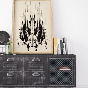 May include: A black and white abstract inkblot print in a wooden frame hanging above a black metal cabinet with drawers and a wire mesh door. The cabinet has books on top, including one titled "History of Home Decor".