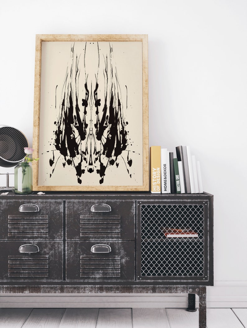 May include: A black and white abstract inkblot print in a wooden frame hanging above a black metal cabinet with drawers and a wire mesh door. The cabinet has books on top, including one titled "History of Home Decor".