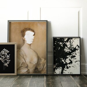 May include: Three framed prints on a wooden floor. The print on the left is a black and white photograph of a flower. The middle print is a portrait of a woman with a white painted over her face. The print on the right is an abstract black and white ink painting.