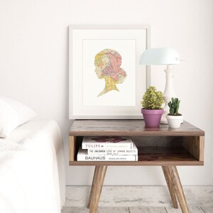 May include: A white framed print with a silhouette of a woman's head made from a vintage map. The print is on a wooden nightstand with two books, a lamp, and two potted plants.