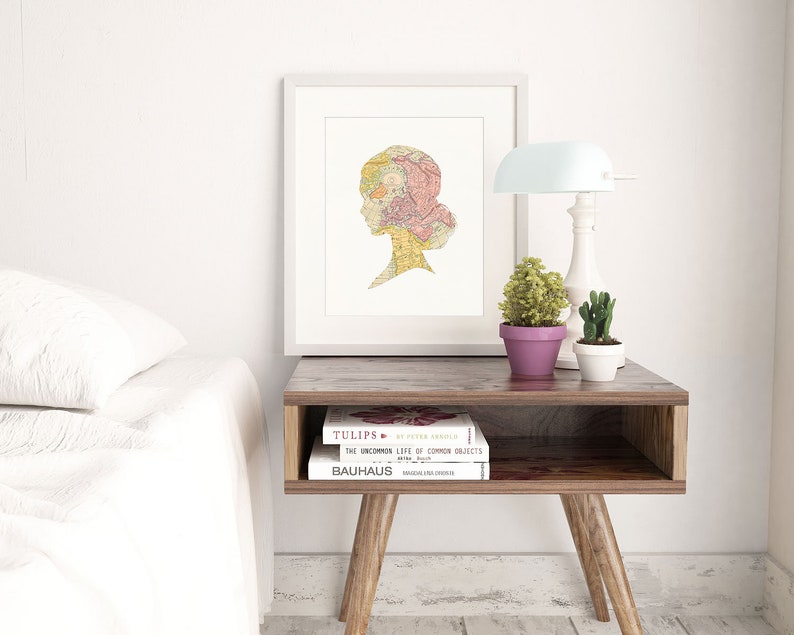May include: A white framed print with a silhouette of a woman's head made from a vintage map. The print is on a wooden nightstand with two books, a lamp, and two potted plants.