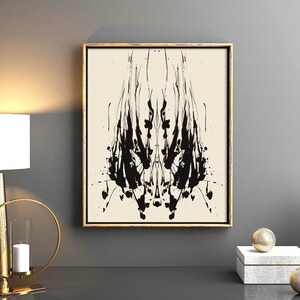 May include: A black and white abstract art print in a gold frame, hanging on a gray wall above a gold metal decorative stand with a candle and two circular metal accents. The stand is on a black surface.