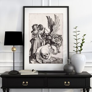 May include: A black and white print of a woman and a man with a skull and a winged creature. The print is framed in a black frame and is hanging on a white wall.
