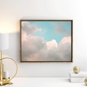 May include: A framed canvas print of a pastel pink and blue sky with fluffy white clouds. The print is hanging on a white wall above a white table with gold decorative objects.