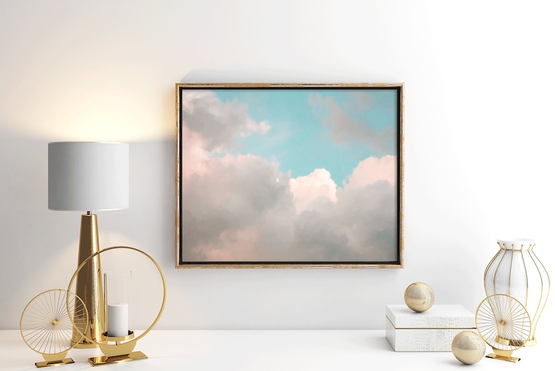 May include: A framed canvas print of a pastel pink and blue sky with fluffy white clouds. The print is hanging on a white wall above a white table with gold decorative objects.