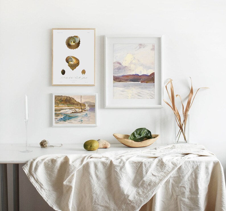May include: Three framed prints on a white wall, a white table with a linen tablecloth, a wooden bowl with a green plant, and a vase with dried branches. The prints are of a seascape, a seashell, and a boat.