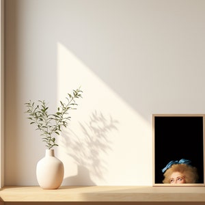 Pu&ograve; includere: Un vaso bianco con una pianta verde si trova su uno scaffale di legno accanto a un quadro incorniciato di una donna con un nastro blu nei capelli. L'immagine &egrave; su uno sfondo nero.