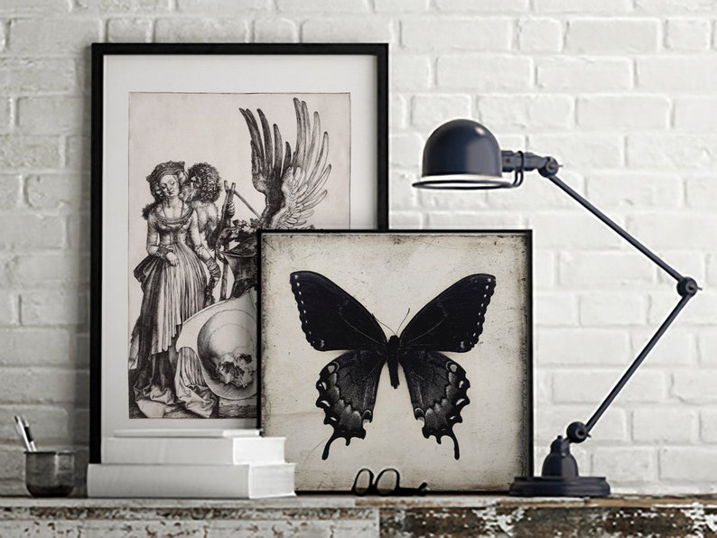May include: Two black and white framed prints on a white brick wall. The print on the left features a black and white engraving of a couple standing next to a skull. The print on the right features a black and white image of a butterfly.