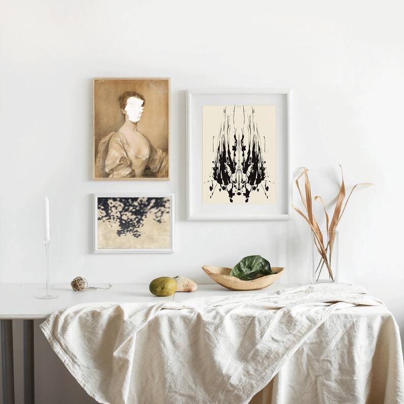 May include: Three framed prints on a white wall above a white linen tablecloth on a table. The print on the left is a portrait of a woman in a white dress. The print in the middle is a black and white abstract design. The print on the right is a photograph of a cloudy sky.
