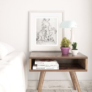 May include: A black and white framed print of a drawing of a horse rearing up with a person standing on its back. The print is on a wooden table with two books and a lamp.
