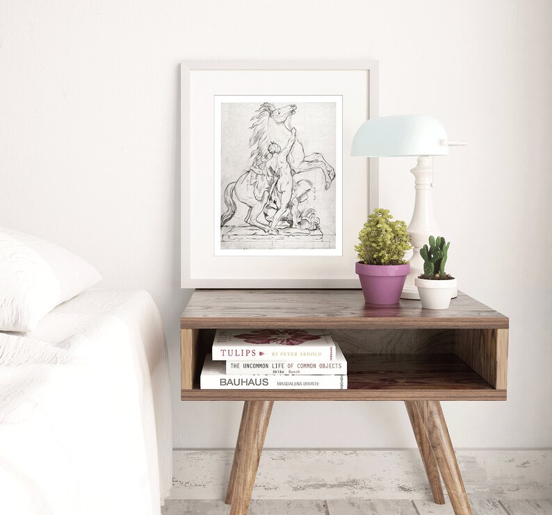 May include: A black and white framed print of a drawing of a horse rearing up with a person standing on its back. The print is on a wooden table with two books and a lamp.