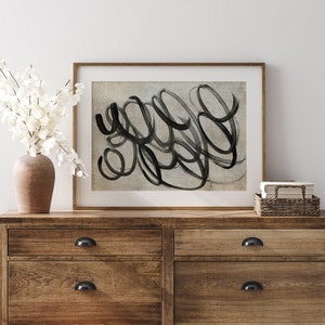 May include: A framed abstract art print with black brushstrokes on a light gray background. The print is hanging on a white wall above a wooden dresser with drawers.