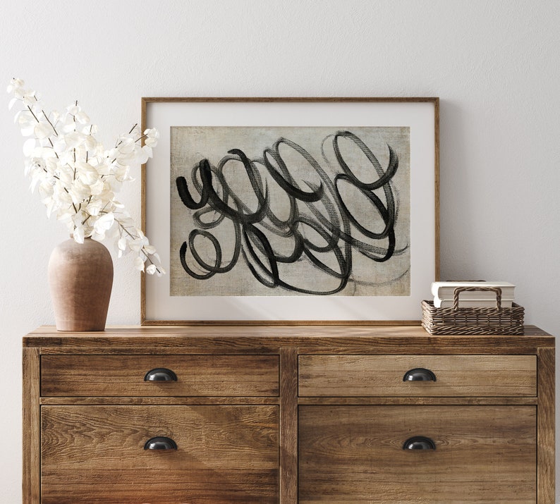 May include: A framed abstract art print with black brushstrokes on a light gray background. The print is hanging on a white wall above a wooden dresser with drawers.