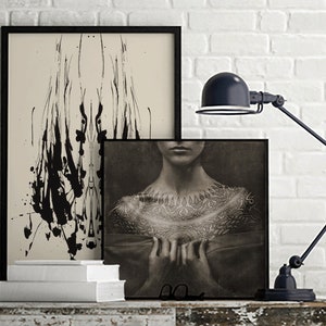 May include: Two framed black and white art prints on a white brick wall. The print on the left features abstract black ink splatter on a cream background. The print on the right features a black and white photo of a person's chest with a detailed tattoo design.
