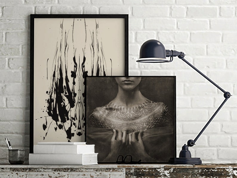 May include: Two framed black and white art prints on a white brick wall. The print on the left features abstract black ink splatter on a cream background. The print on the right features a black and white photo of a person's chest with a detailed tattoo design.