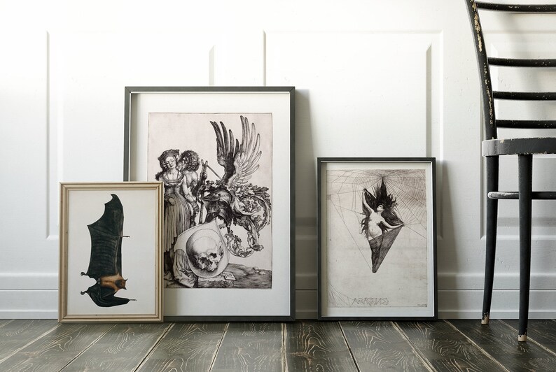 May include: Three framed black and white prints on a white wall with a black wooden chair in the corner. The prints are of a bat, a couple with a skull and a woman in a spider web.