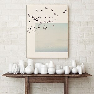 May include: A framed print of a flock of birds flying over a body of water. The print is hanging on a white brick wall above a wooden console table with white ceramic vases.