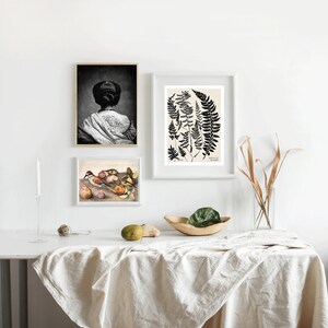 May include: Three framed prints on a white wall above a table with a white tablecloth. The prints are a black and white photo of a woman with her back to the camera, a black and white illustration of ferns, and a painting of birds and fruit.