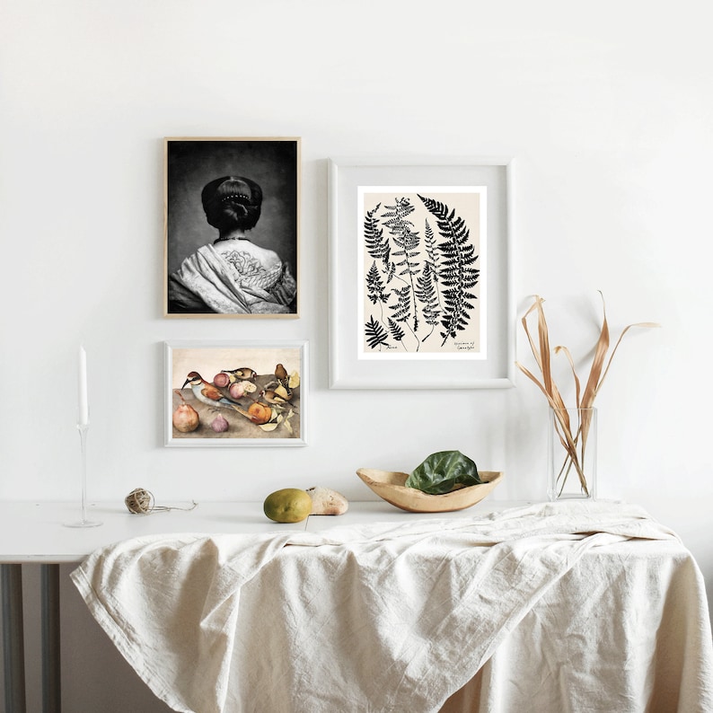 May include: Three framed prints on a white wall above a table with a white tablecloth. The prints are a black and white photo of a woman with her back to the camera, a black and white illustration of ferns, and a painting of birds and fruit.
