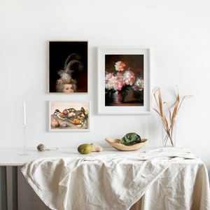 May include: Three framed art prints on a white wall. The prints feature a portrait of a woman, a still life of flowers, and a still life of fruit and birds. A white table with a linen tablecloth is in the foreground.