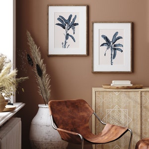 May include: Two framed prints of blue banana leaf illustrations on a light brown wall. The prints are hanging above a wooden cabinet with a white geometric pattern. A brown leather chair sits in front of the cabinet.