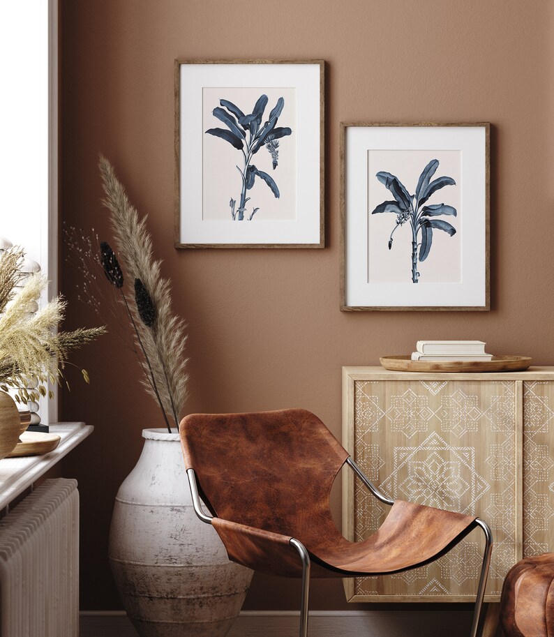 May include: Two framed prints of blue banana leaf illustrations on a light brown wall. The prints are hanging above a wooden cabinet with a white geometric pattern. A brown leather chair sits in front of the cabinet.