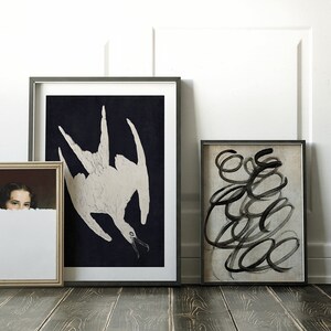 May include: Three framed pictures on a wooden floor. The picture on the left is a portrait of a woman with dark hair. The middle picture is a black and white illustration of a bird. The picture on the right is an abstract black and white painting.
