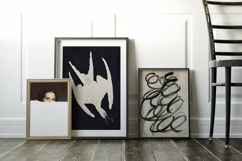 May include: Three framed pictures on a wooden floor. The picture on the left is a portrait of a woman with dark hair. The middle picture is a black and white illustration of a bird. The picture on the right is an abstract black and white painting.