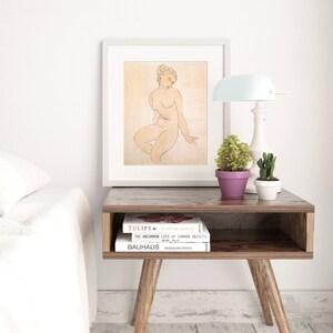 May include: A white framed print of a line drawing of a nude woman sitting with her head tilted down. The print is titled "Tulips" by Peter Arnold. The print is on a wooden end table with two books stacked on top of each other. The books are titled "Tulips" by Peter Arnold and "The Uncommon Life of Common Objects" by Arthur Waugh. The bottom book is titled "Bauhaus" by Magdalena Droste.
