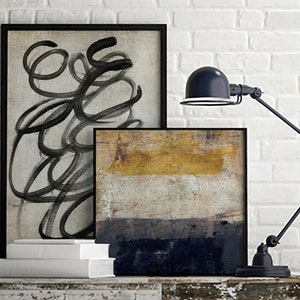 May include: Two framed abstract paintings on a white brick wall with a black desk lamp. The painting on the left is black and white with abstract lines. The painting on the right is a multi-colored abstract painting with shades of yellow, white, and blue.