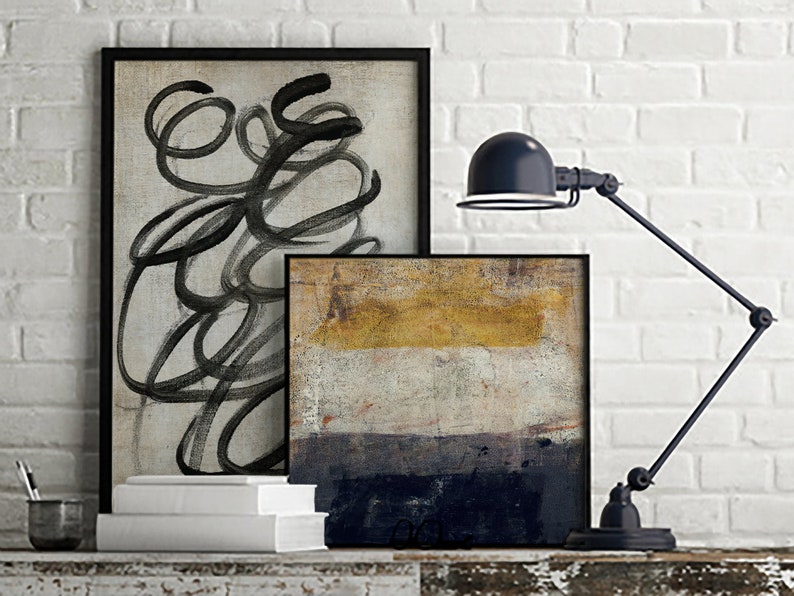 May include: Two framed abstract paintings on a white brick wall with a black desk lamp. The painting on the left is black and white with abstract lines. The painting on the right is a multi-colored abstract painting with shades of yellow, white, and blue.
