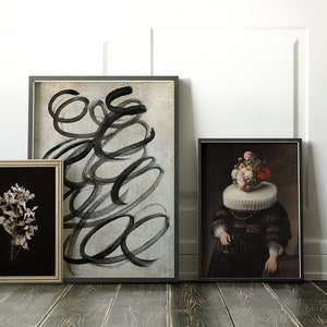 May include: Three framed art prints on a wood floor. The print on the left is a black and white photo of dried flowers. The middle print is a black and white abstract painting with swirling lines. The print on the right is a painting of a person with a bouquet of flowers on their head.