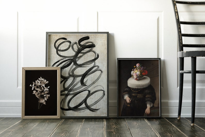 May include: Three framed art prints on a wood floor. The print on the left is a black and white photo of dried flowers. The middle print is a black and white abstract painting with swirling lines. The print on the right is a painting of a person with a bouquet of flowers on their head.
