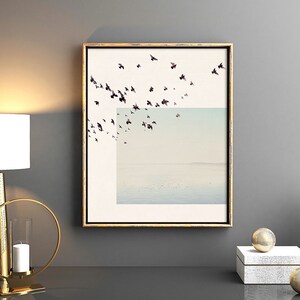 May include: A framed print of a flock of birds flying over a body of water. The birds are black and the water is a light blue color. The frame is gold.