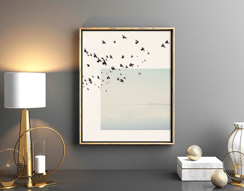 May include: A framed print of a flock of birds flying over a body of water. The birds are black and the water is a light blue color. The frame is gold.