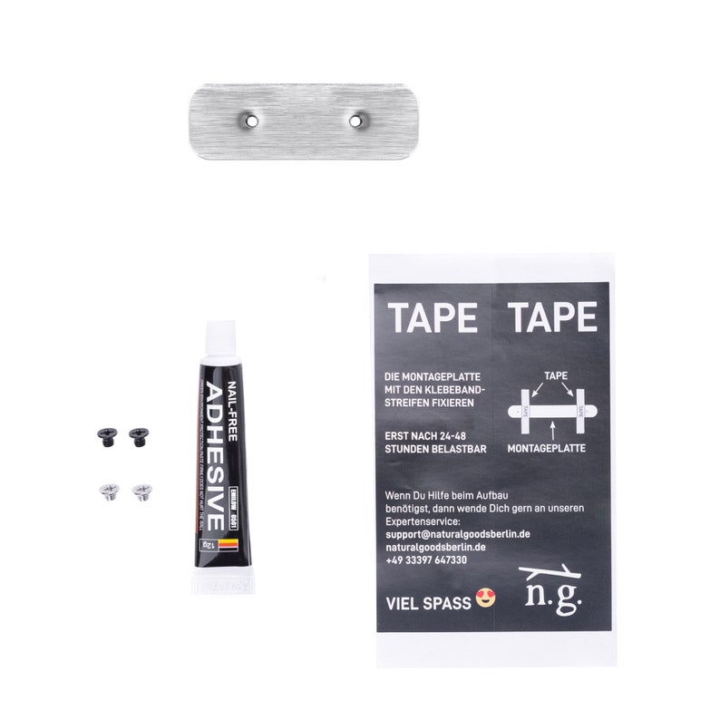 Op de afbeelding: Een zilveren metalen montageplaat met twee gaten voor schroeven. De plaat wordt geleverd met een tube nagelvrije lijm, twee schroeven en instructies voor montage met tape. De instructies zijn in het Duits en bevatten de tekst "TAPE TAPE" en "VIEL SPASS n.g."