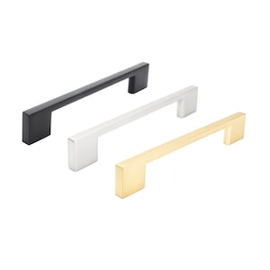 Può includere: Tre maniglie per mobili in diverse finiture metalliche: nero opaco, argento spazzolato e oro spazzolato. Ogni maniglia ha un design a barra rettangolare con un'impugnatura quadrata. Le maniglie sono disposte diagonalmente su uno sfondo bianco.