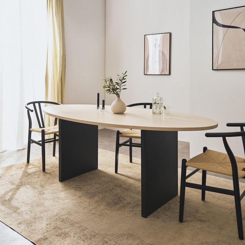 Op de afbeelding: Een ovale eettafel met een licht houten blad en een zwarte basis. De tafel is omringd door zwarte stoelen met geweven zittingen. Een vaas met groen en andere decoratieve items staan op de tafel.