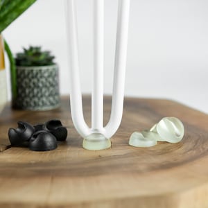 Może przedstawiać: Białe metalowe nogi meblowe z przezroczystymi plastikowymi osłonami na podłogę. Na zdjęciu znajdują się również czarne plastikowe osłony na podłogę.