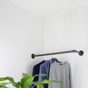 Könnte beinhalten: Ein schwarzer Metallrohr-Kleiderständer, der an einer weißen Wand montiert ist, mit drei Kleiderbügeln, an denen ein blau-grauer Pullover, ein blaues Hemd mit Knöpfen und ein grauer Pullover hängen. Im Vordergrund befindet sich eine grüne Pflanze.