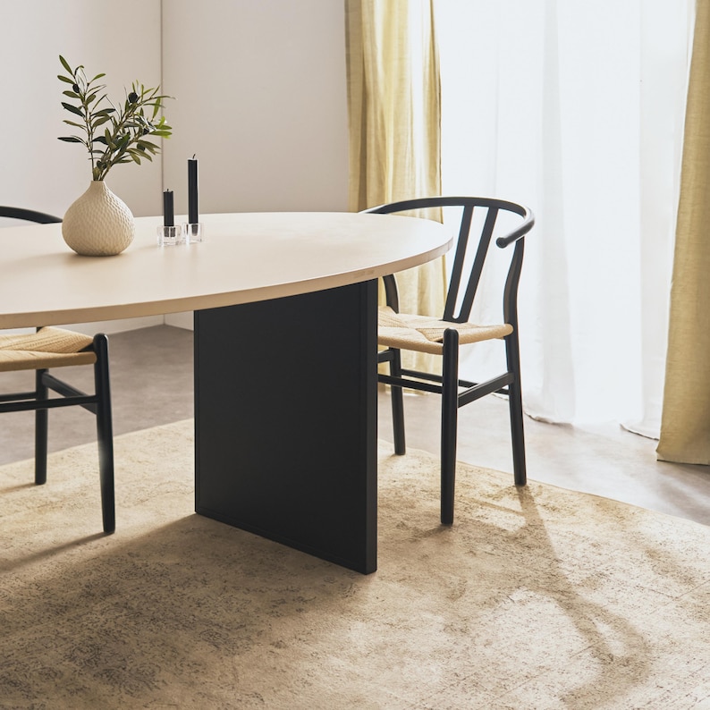 Op de afbeelding: Een moderne eethoek met een ovale tafel van licht hout en een zwarte voet. Twee zwarte stoelen met geweven zittingen staan rond de tafel. Een beige tapijt en een vaas met groen maken de sc&egrave;ne compleet.