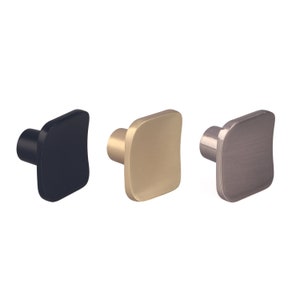 Op de afbeelding: Drie kastknoppen in verschillende afwerkingen: zwart, goud en geborsteld nikkel. Elke knop heeft een vierkant oppervlak met afgeronde randen en een cilindrische basis. Geschikt voor laden en kasten.