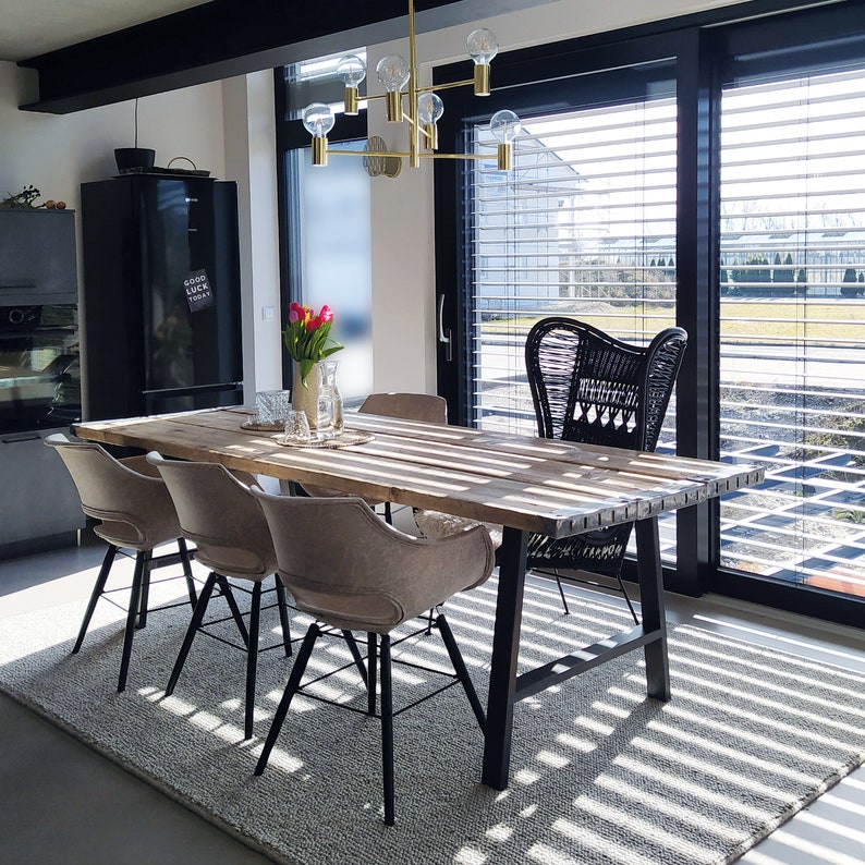 Puede incluir: Una mesa de comedor de madera con patas de metal negro y cuatro sillas. La mesa tiene un aspecto r&uacute;stico de madera recuperada. Las sillas est&aacute;n tapizadas en tela gris claro. La mesa est&aacute; puesta con un jarr&oacute;n de flores rosas y algunos otros elementos decorativos. La mesa est&aacute; en un comedor moderno con grandes ventanales y una nevera negra.