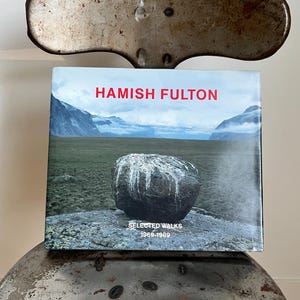 May include: A book titled "Hamish Fulton: Selected Walks 1969-1989" with a photo of a large, grey rock on a green field with mountains in the background.
