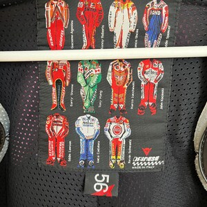 May include: A black leather jacket with a Dainese logo and a patch featuring a grid of motorcycle racers in their racing suits. The patch includes the names of the racers, including Giacomo Agostini, Kenny Roberts, and Eddie Lawson. The jacket has a mesh lining and a 56 logo on the bottom.
