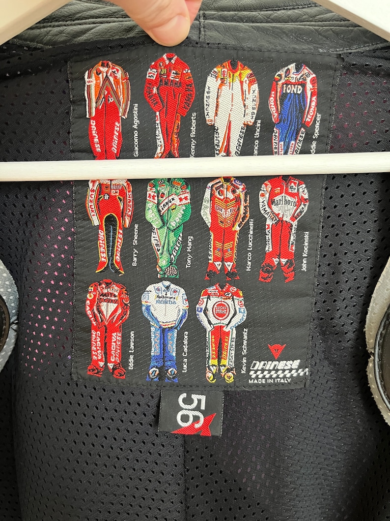 May include: A black leather jacket with a Dainese logo and a patch featuring a grid of motorcycle racers in their racing suits. The patch includes the names of the racers, including Giacomo Agostini, Kenny Roberts, and Eddie Lawson. The jacket has a mesh lining and a 56 logo on the bottom.